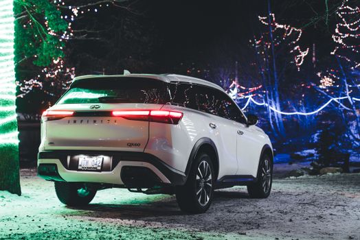 Elegant white Infiniti QX60 SUV parked amidst vibrant Christmas lights, creating a festive twilight ambiance.