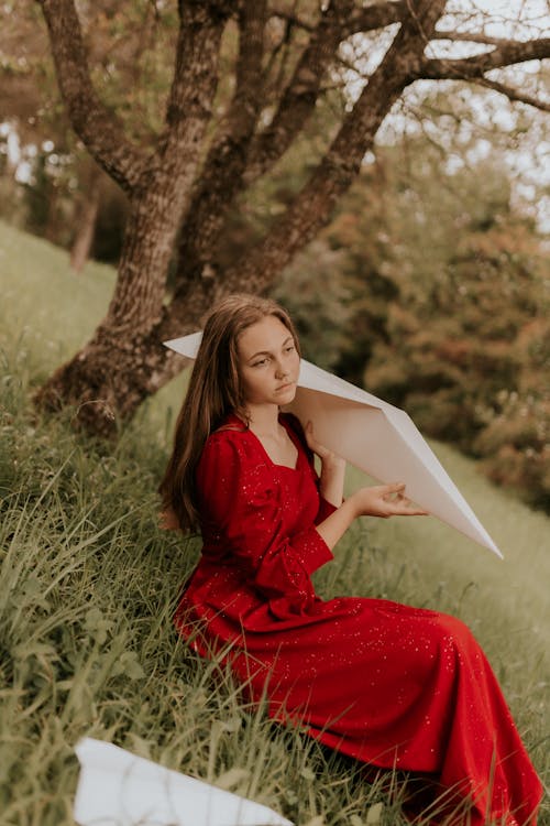 Woman in Red Dress Sitting with Paper Plane on Grass