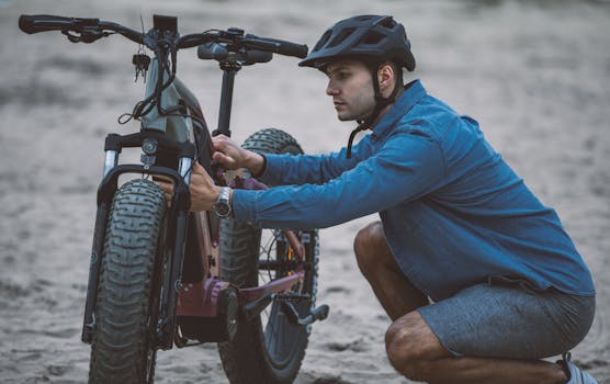 A man in a blue shirt and helmet fixing an electric bicycle on sandy terrain.