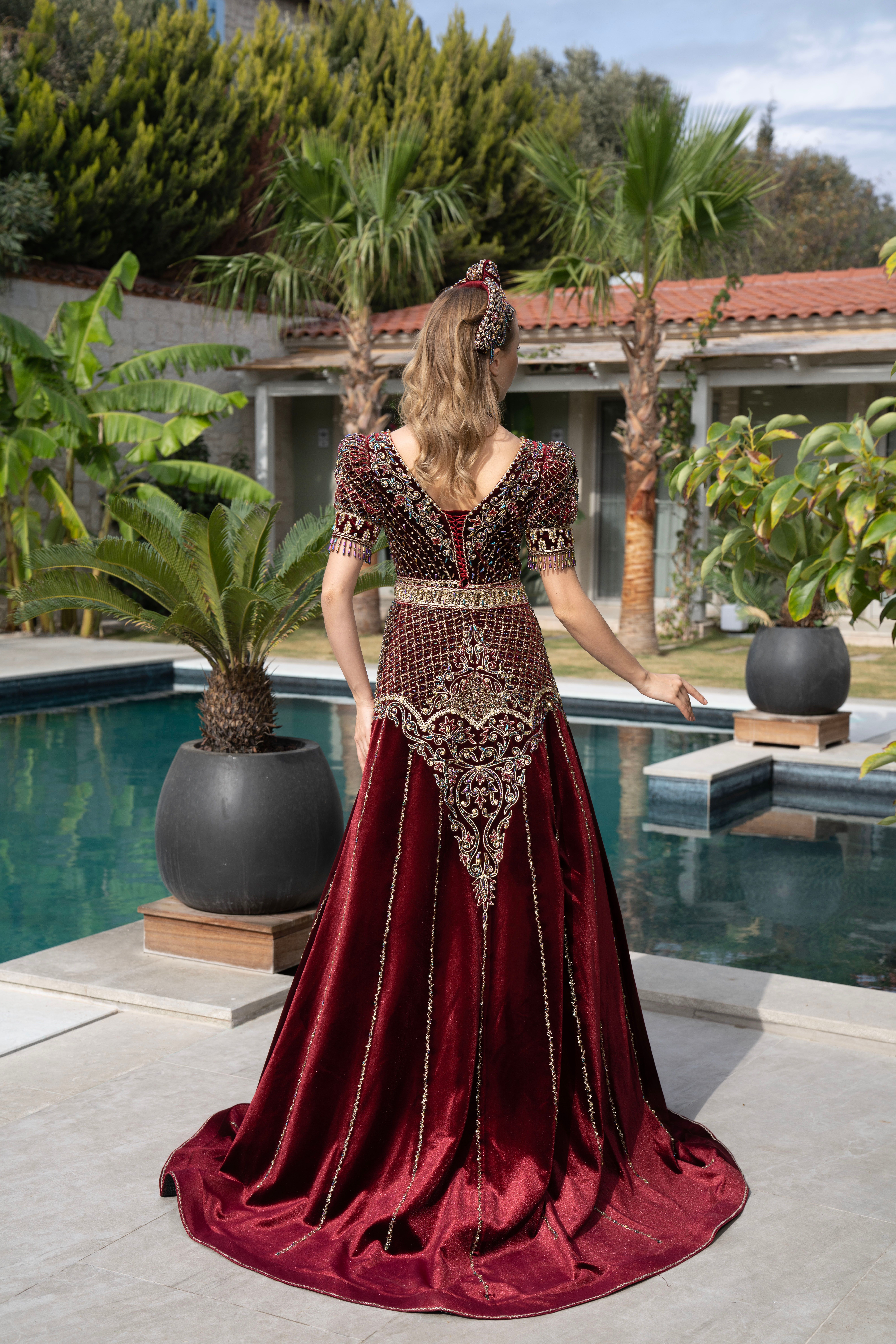 A woman in a luxurious maroon traditional dress stands by a pool, showcasing intricate patterns.