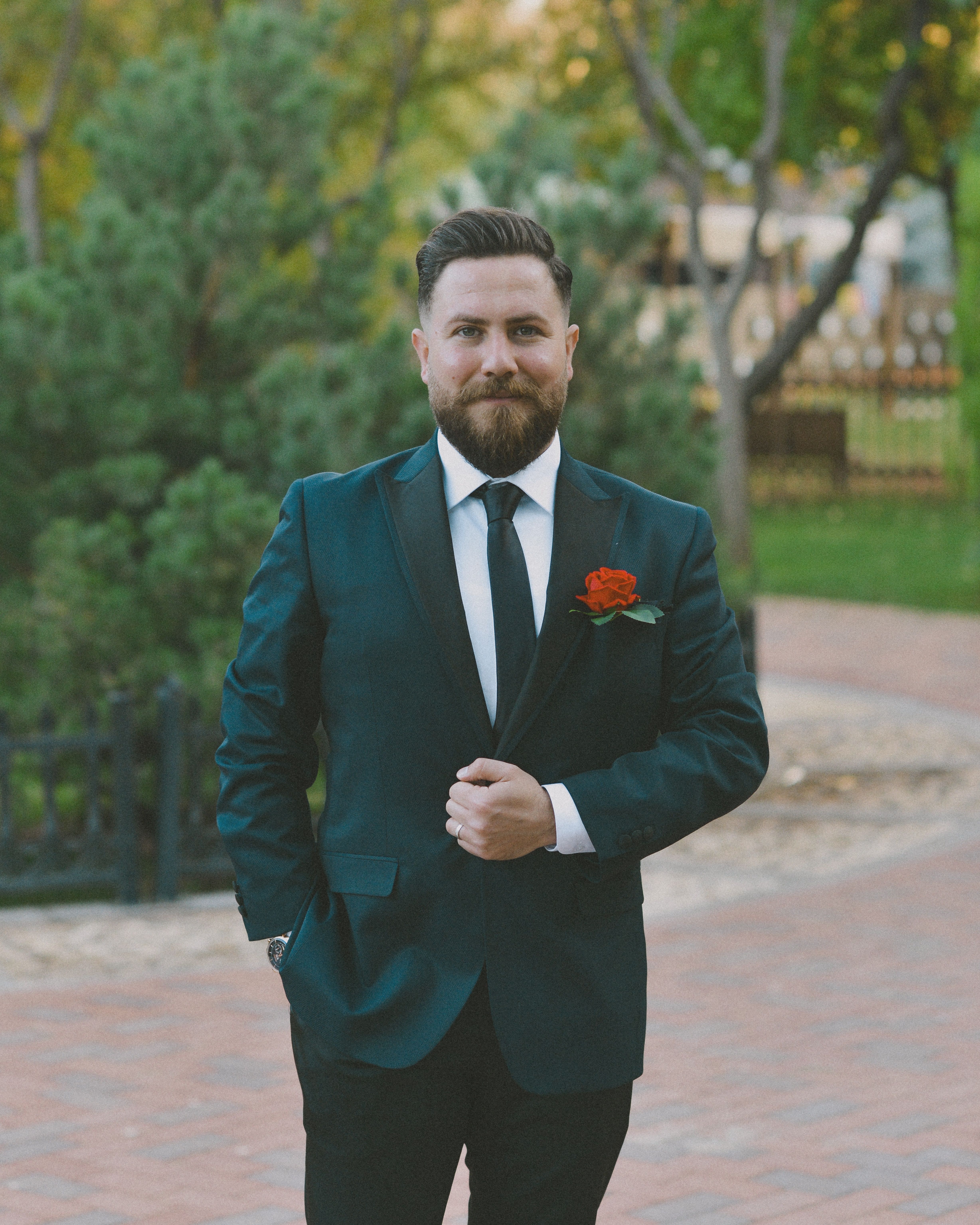 A bearded man in a black suit with a red rose boutonnière, posing outdoors on a sunny day.