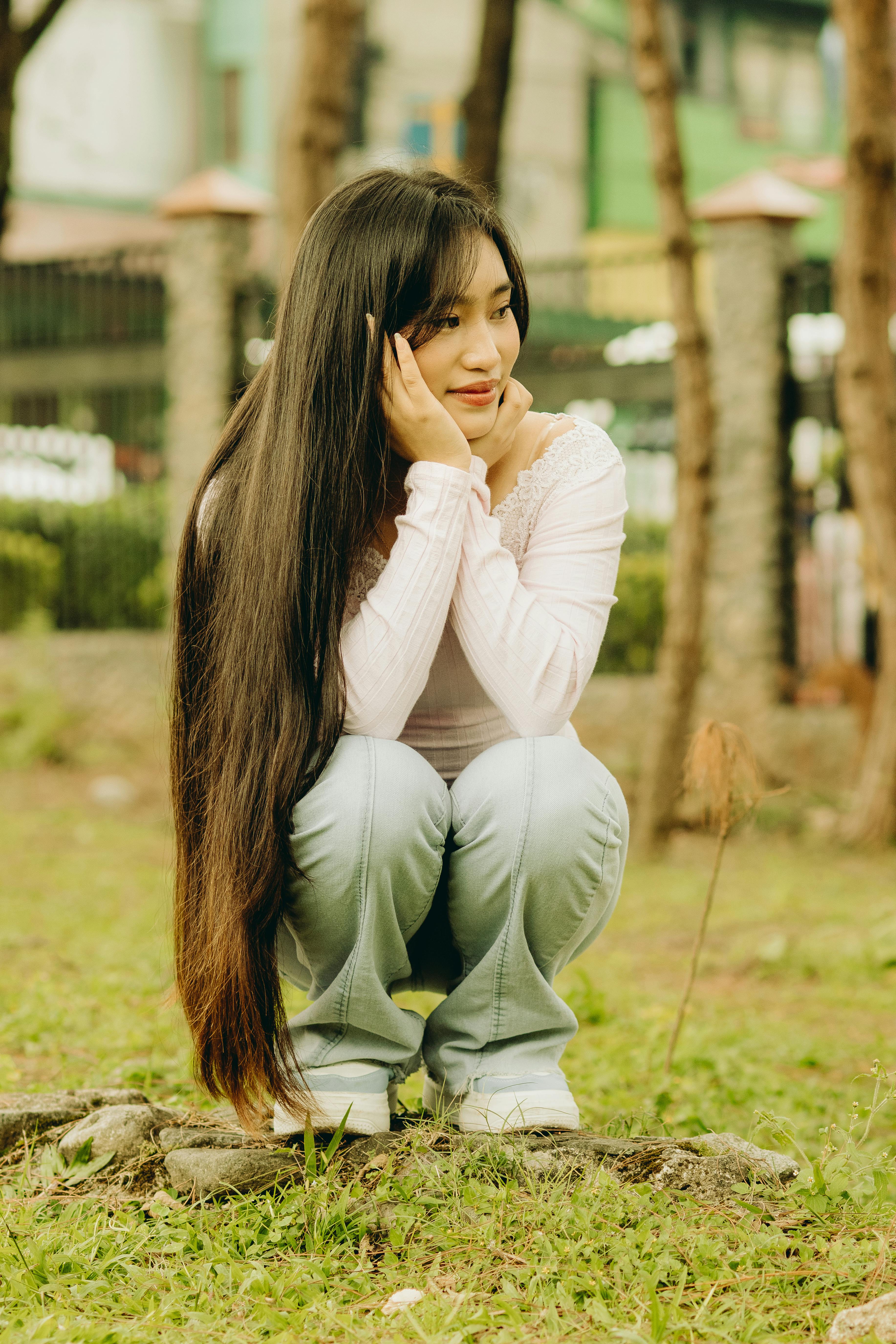 Young Asian woman with long hair squatting in a park, looking thoughtful and serene.