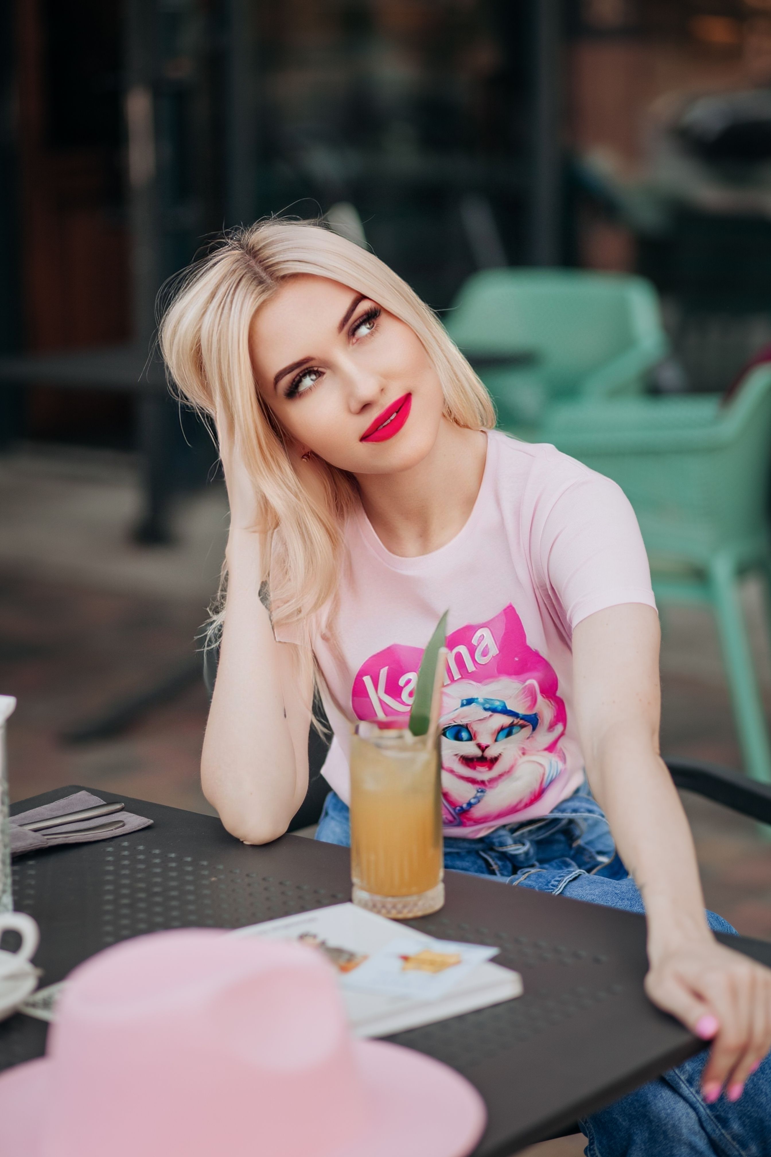 Blonde woman in casual pink attire relaxes with a drink, enjoying leisure time outdoors.