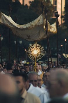 A solemn religious procession featuring a golden monstrance held aloft under a canopy at dusk.