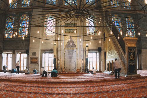 Free A serene mosque interior with people praying, featuring ornate windows and chandeliers. Stock Photo