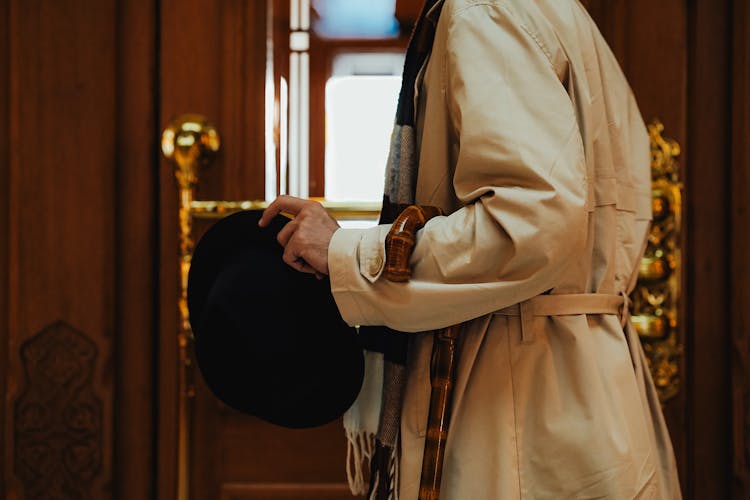 Man In A Beige Trench Coat With A Cane Holding A Black Hat In His Hand Leaving The Hotel