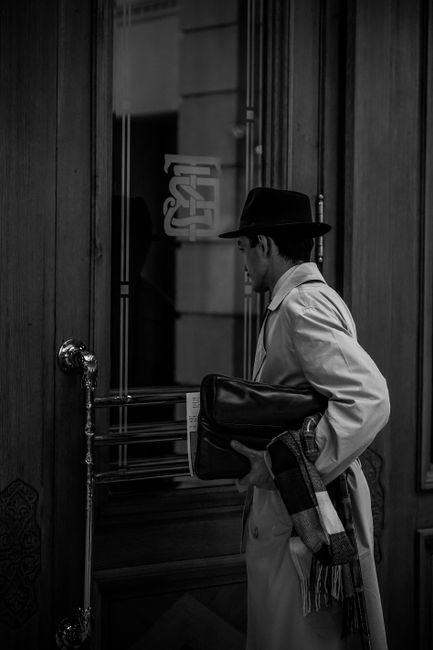 A black and white image of a man in a trench coat holding a bag at a door, symbolizing urban fashion.