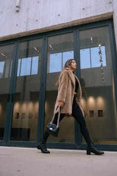 Fashion-forward woman in a fur coat and boots walking past modern architecture with a handbag.