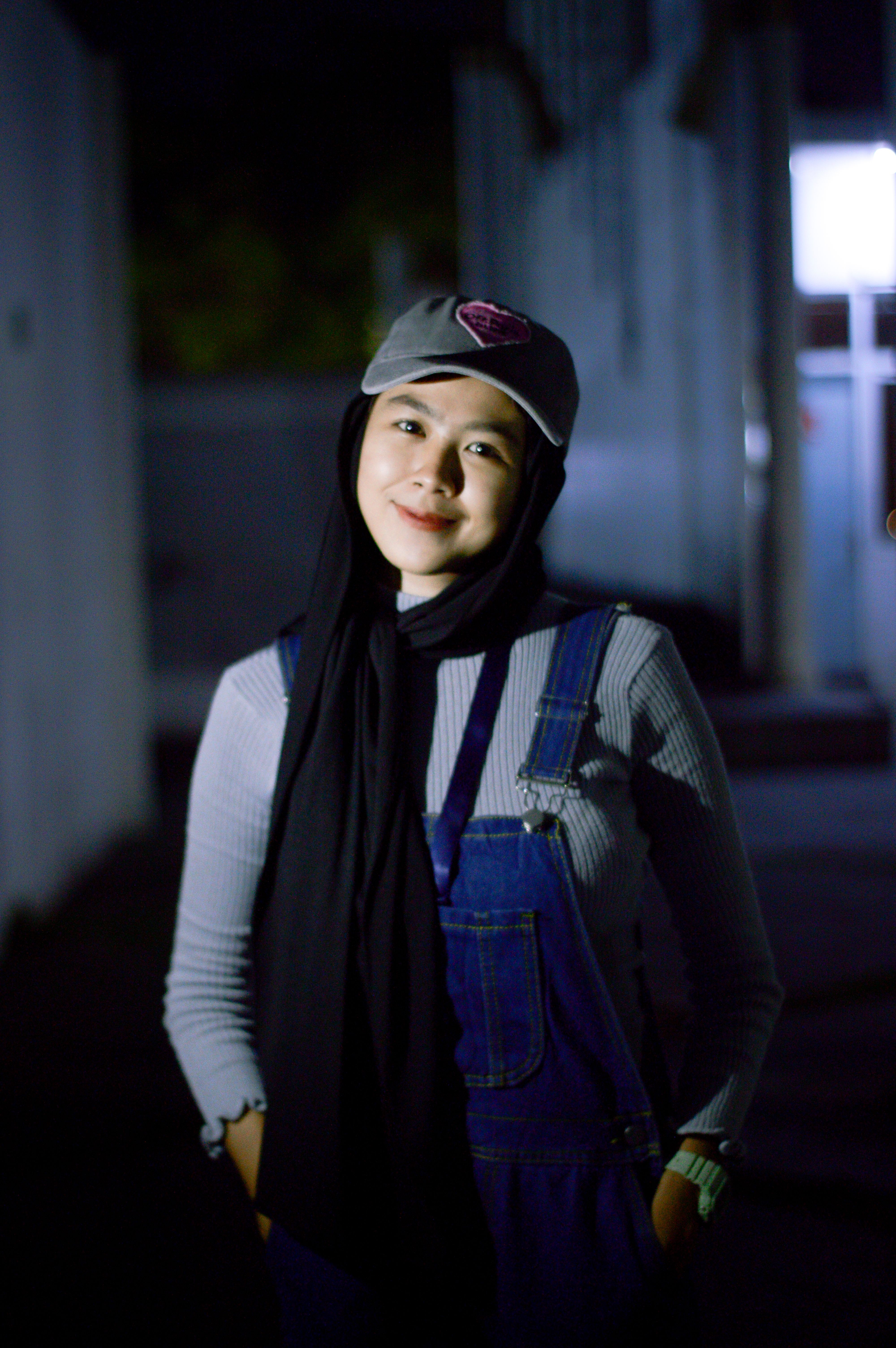 Smiling woman in denim overalls and hat, captured in a nighttime urban setting.