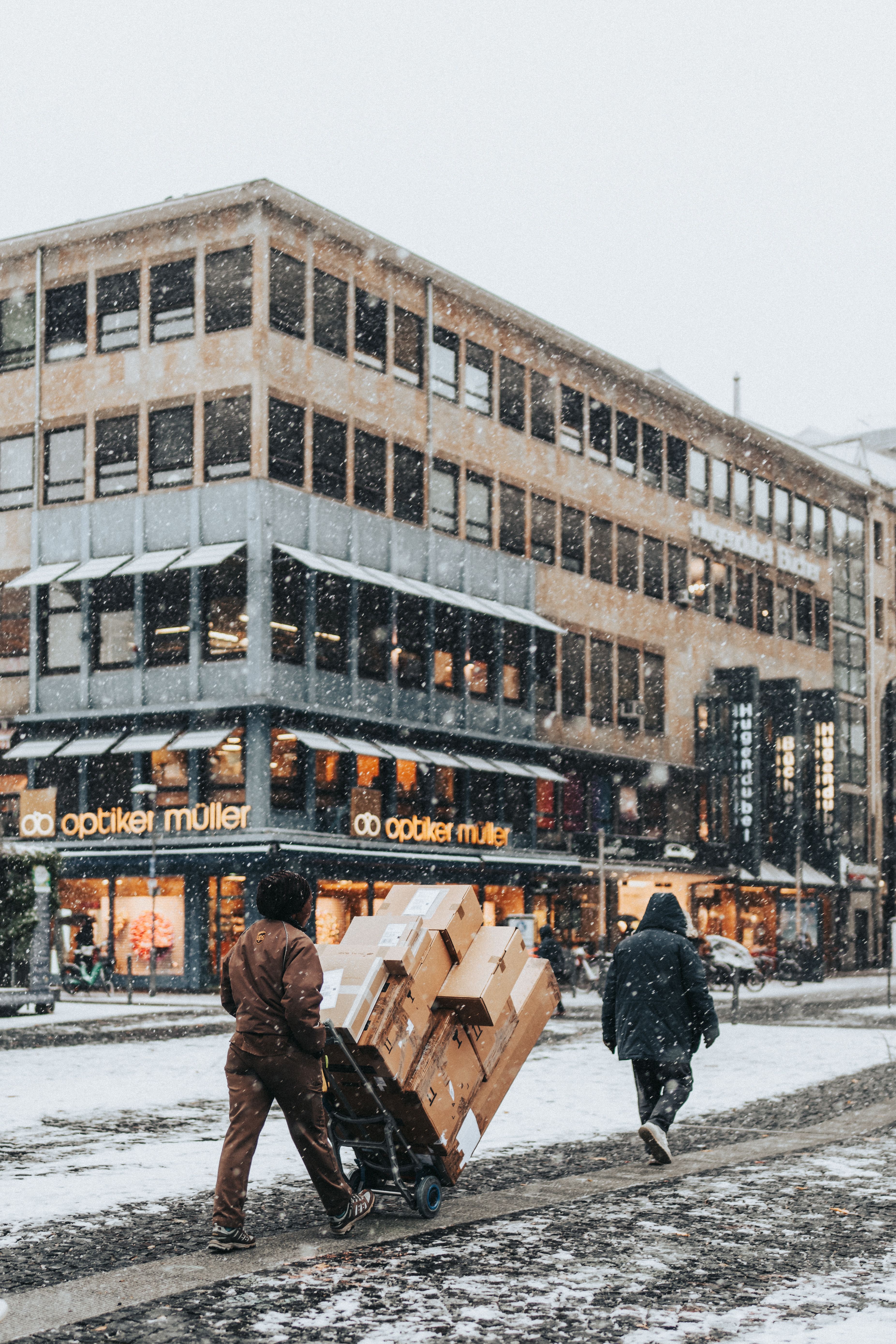 A snowy winter street scene with people walking and a commercial building in the city.