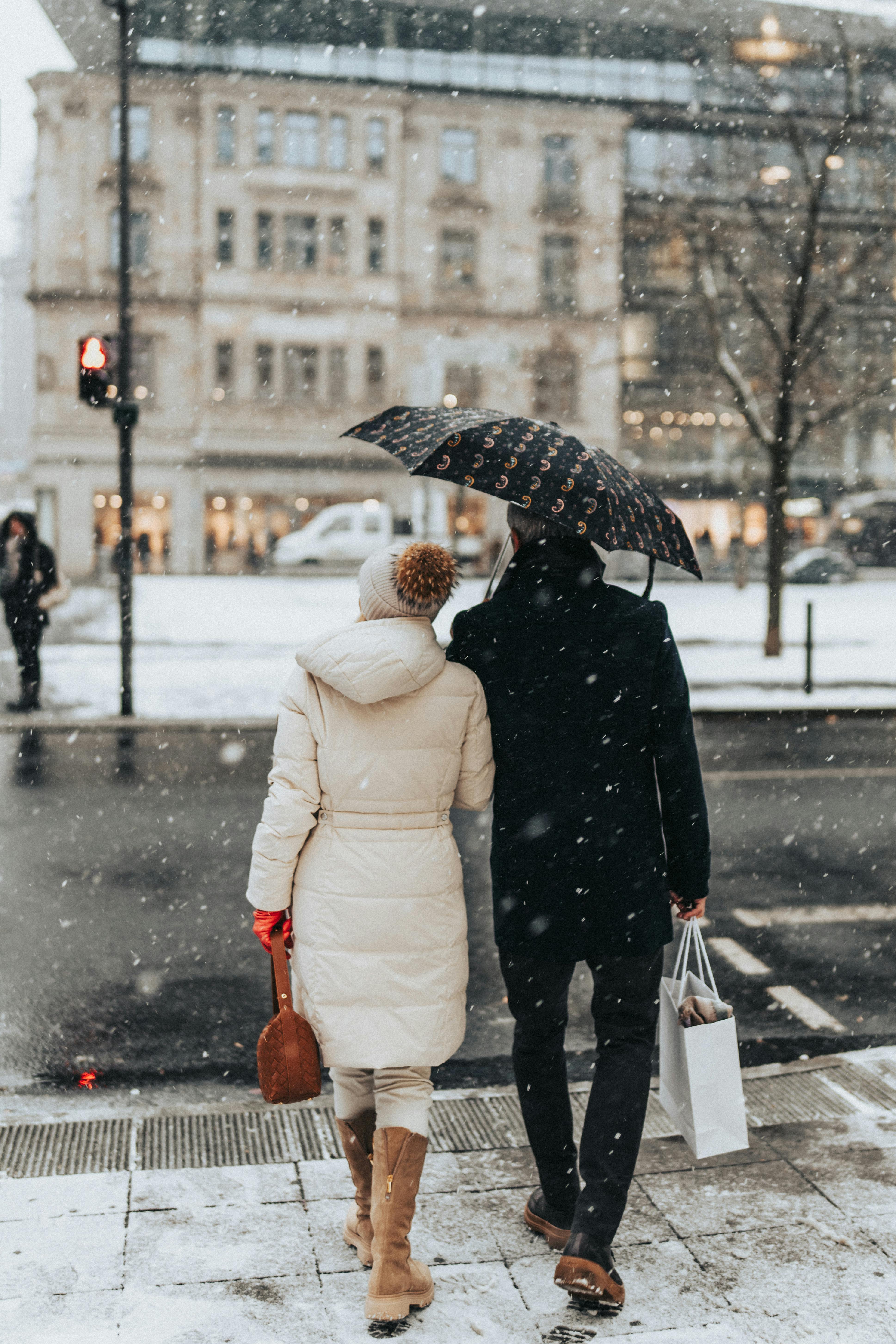 Back view of a couple walking on a snowy city street holding an umbrella, showcasing urban winter fashion.