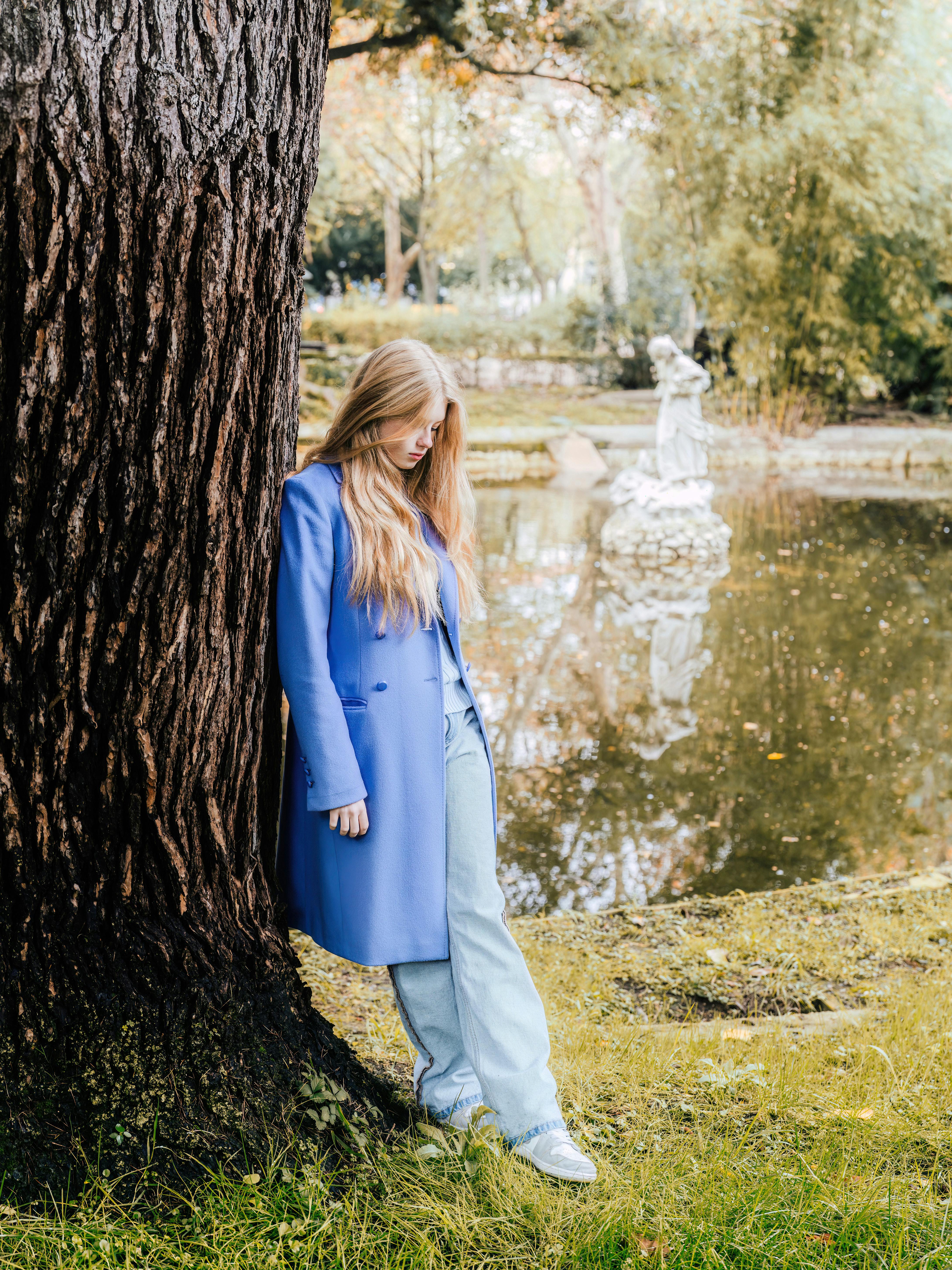 A woman wearing a blue coat leans against a tree by a lake in a Lisbon park during autumn.
