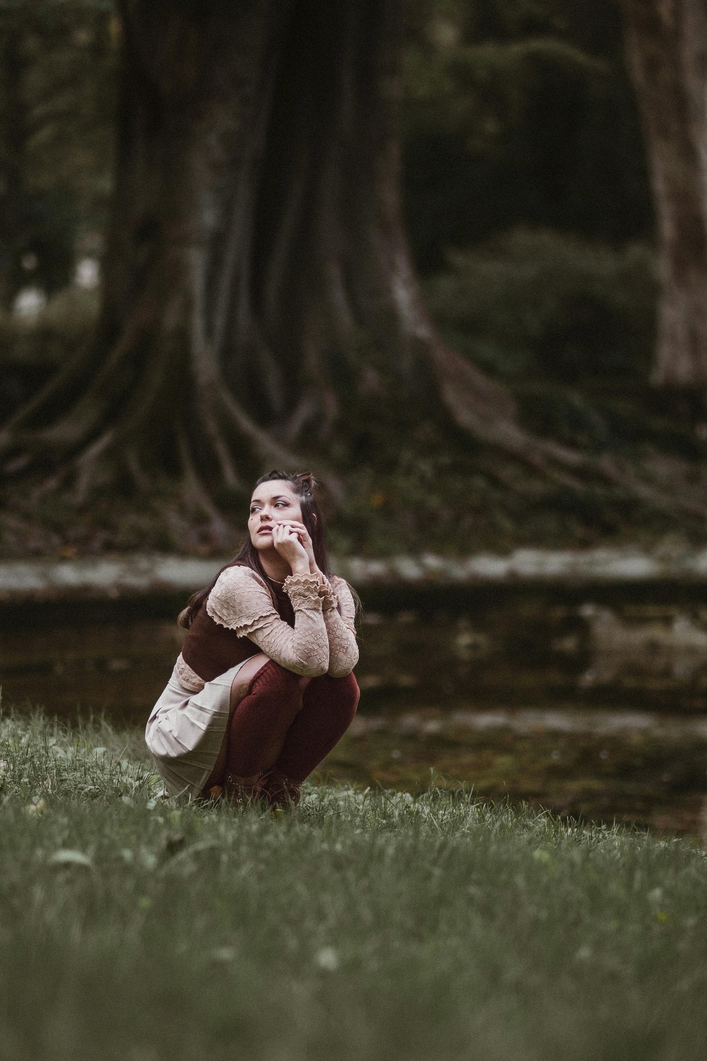 Free A young woman in a park exhibits a contemplative pose amidst lush trees and grass. Stock Photo
