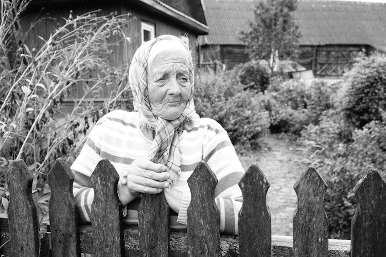 Black And White Photo Of An Elderly Woman Standing By A Wooden Fence 