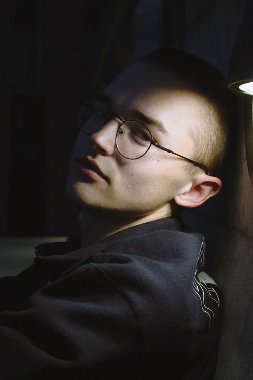 Free A young man in casual clothing and eyeglasses sits in shadowed light against a wall. Stock Photo