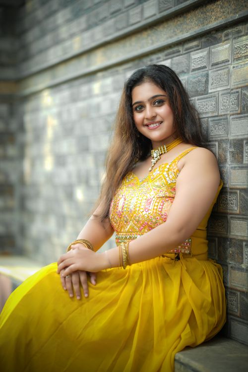 Free A woman in a vibrant yellow traditional dress smiles while posing against a textured brick wall outdoors. Stock Photo