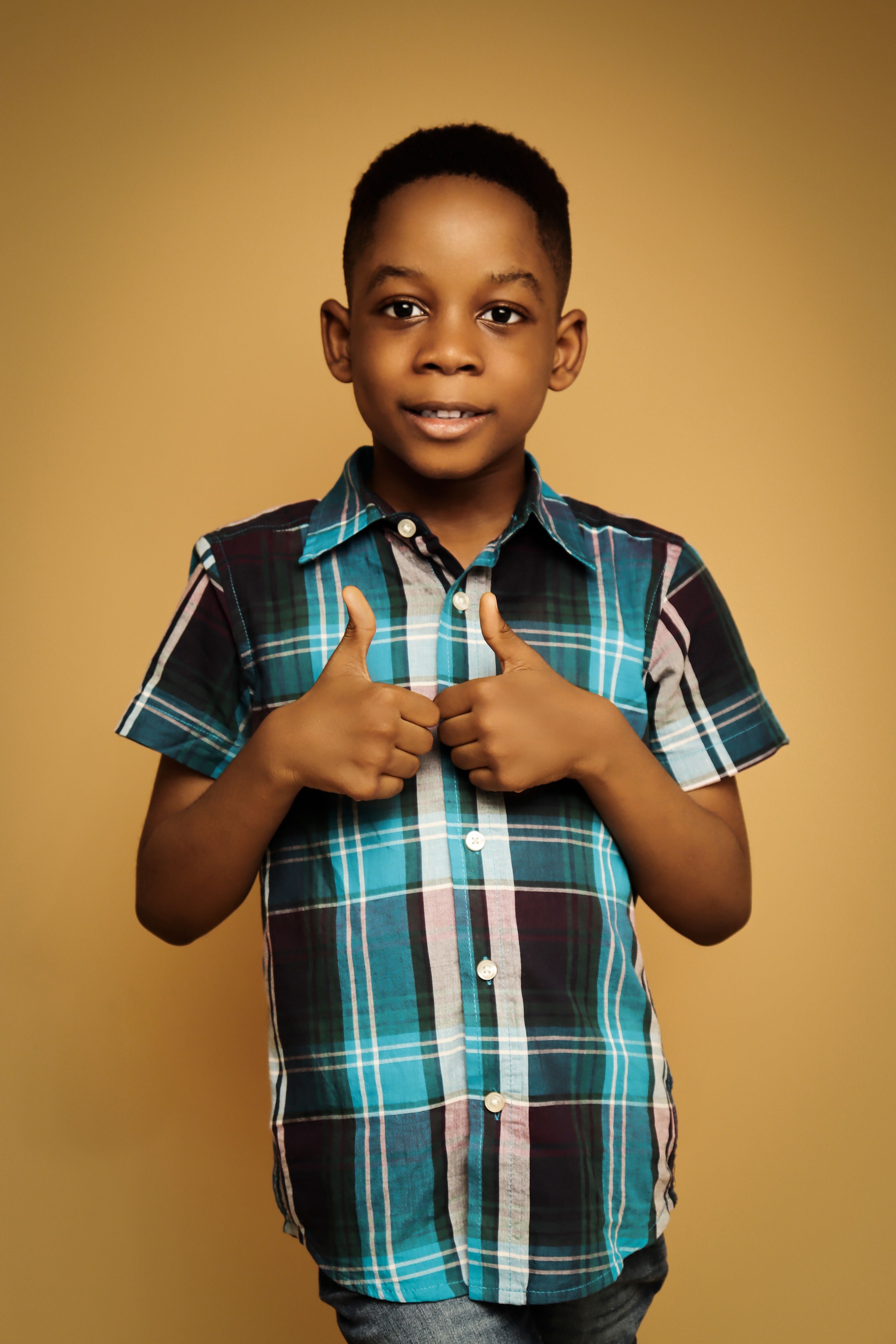 A smiling boy in a colorful plaid shirt giving thumbs up against a yellow background.