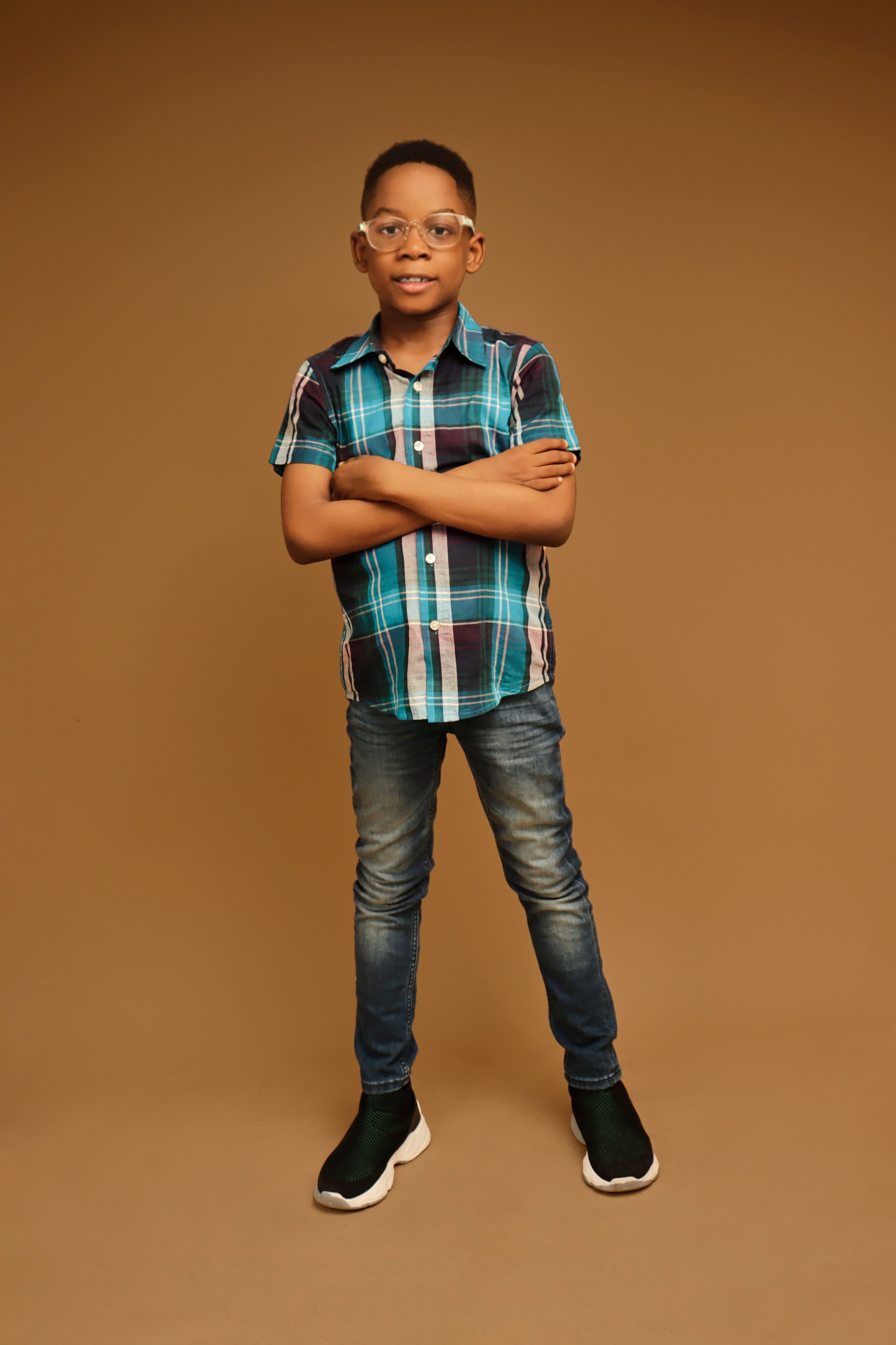 Portrait of a young boy wearing eyeglasses and a checkered shirt with crossed arms. Studio setting.