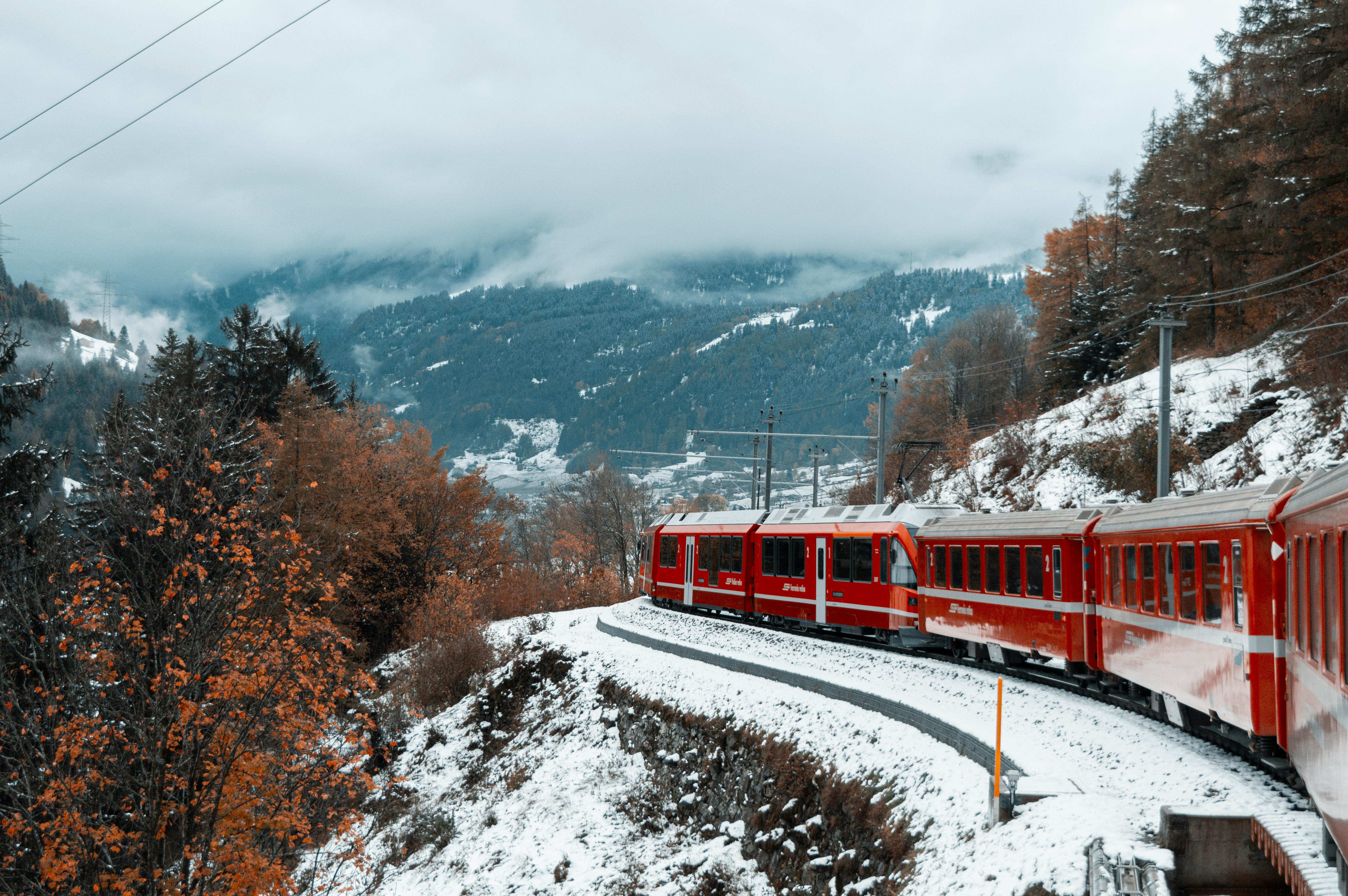 Red train travels through snowy mountains in winter landscape with fog and fall foliage.