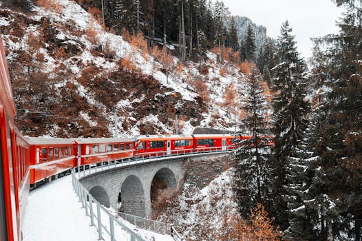 A vibrant red train crosses a snowy coniferous landscape on a scenic mountain bridge in winter.