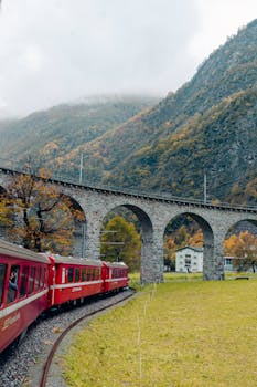 Red train passing under stone arches in a mountainous autumn landscape.