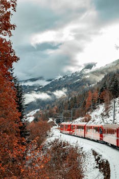 A vibrant red train travels through a snow-covered, mountainous valley surrounded by coniferous trees.