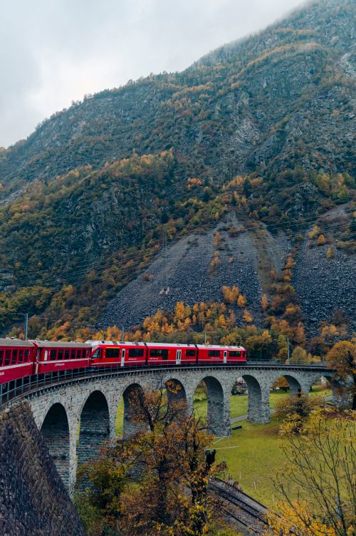 Free Red train traversing a scenic arch bridge in the Swiss Alps during autumn. Stock Photo
