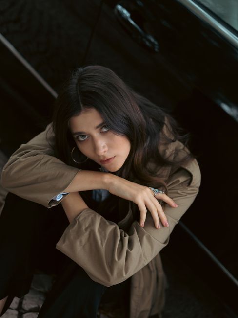 Young woman with brown hair poses stylishly next to a black car in an urban environment.