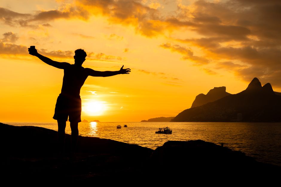 A man celebrates freedom as he stands on a rock, silhouetted against a stunning seaside sunset.
