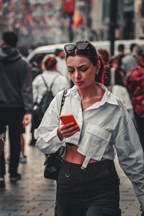 Free A young woman walking through a busy urban street, focused on her smartphone. Stock Photo