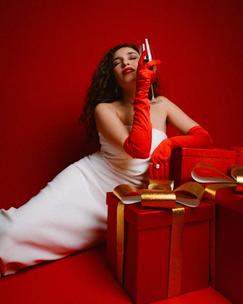 Woman in a White Dress Leaning on Christmas Presents and Drinking Champagne 