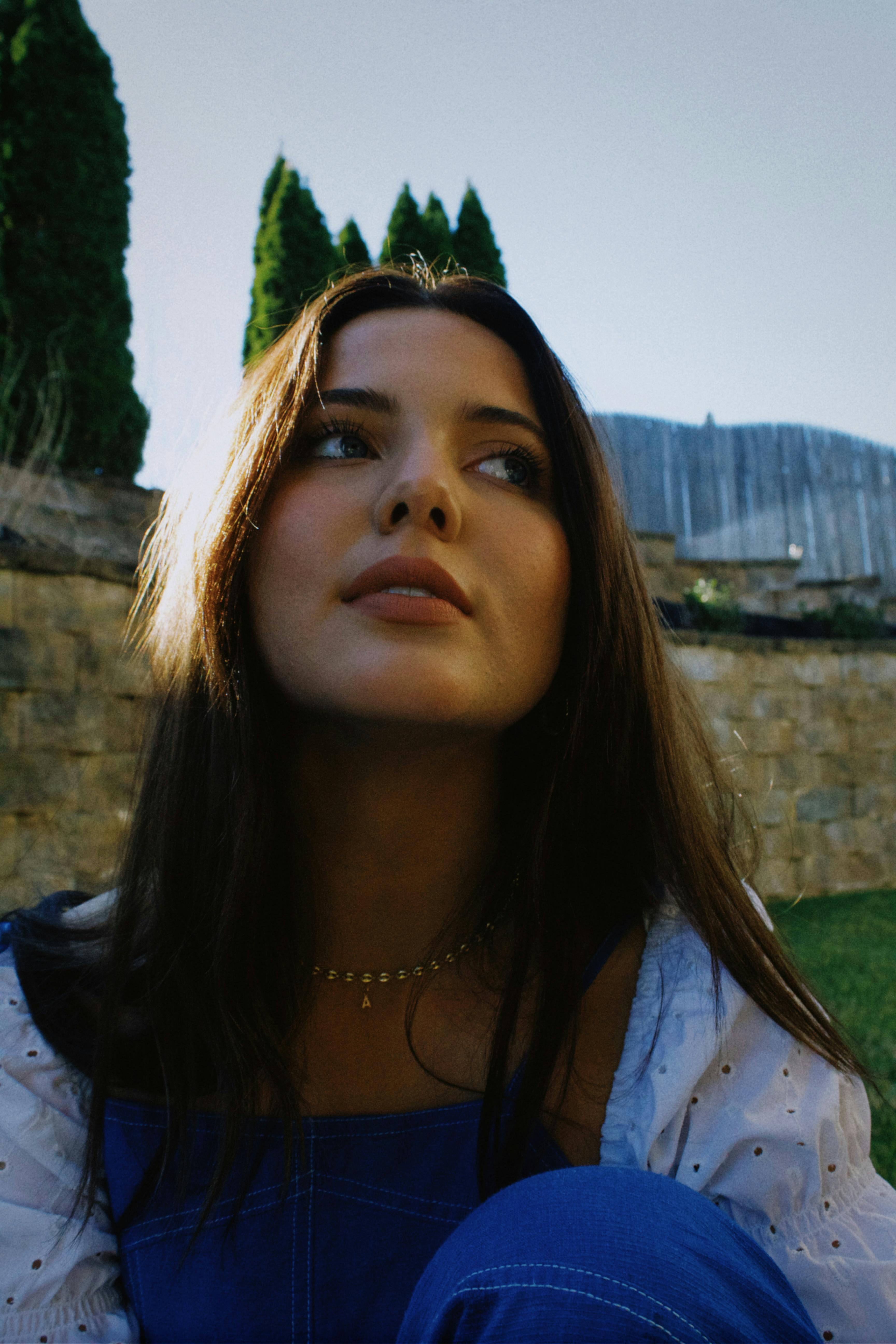 A young woman with brunette hair looks upward in an outdoor setting, showcasing a thoughtful expression.