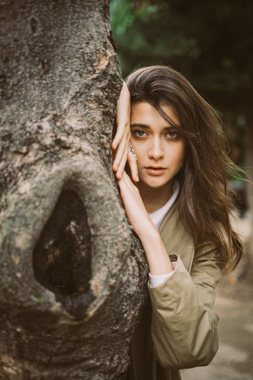 Beautiful Young Brunette Leaning Out From Behind a Tree Trunk