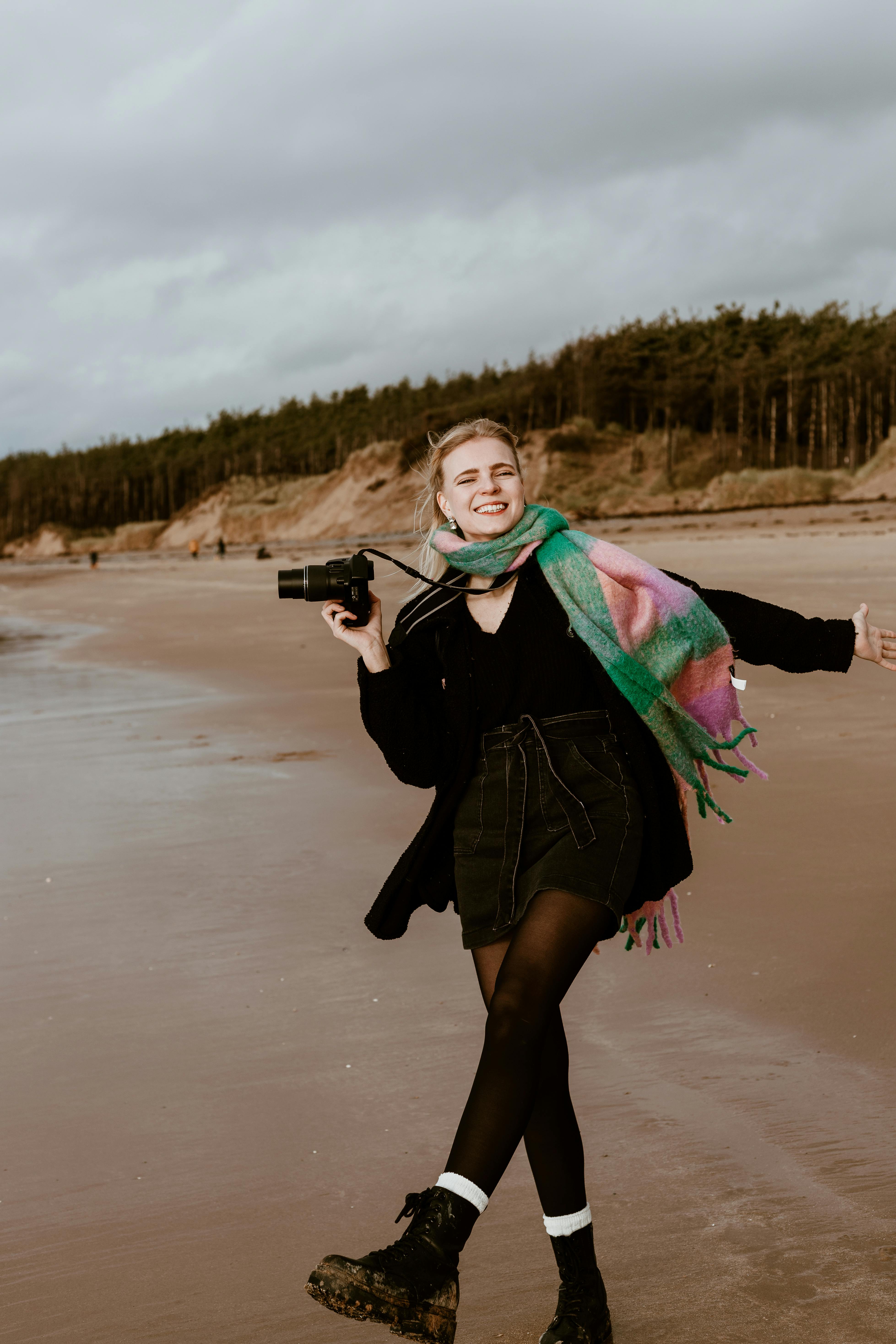 Smiling woman with camera enjoying a windy beach day, showcasing fashion and joy.