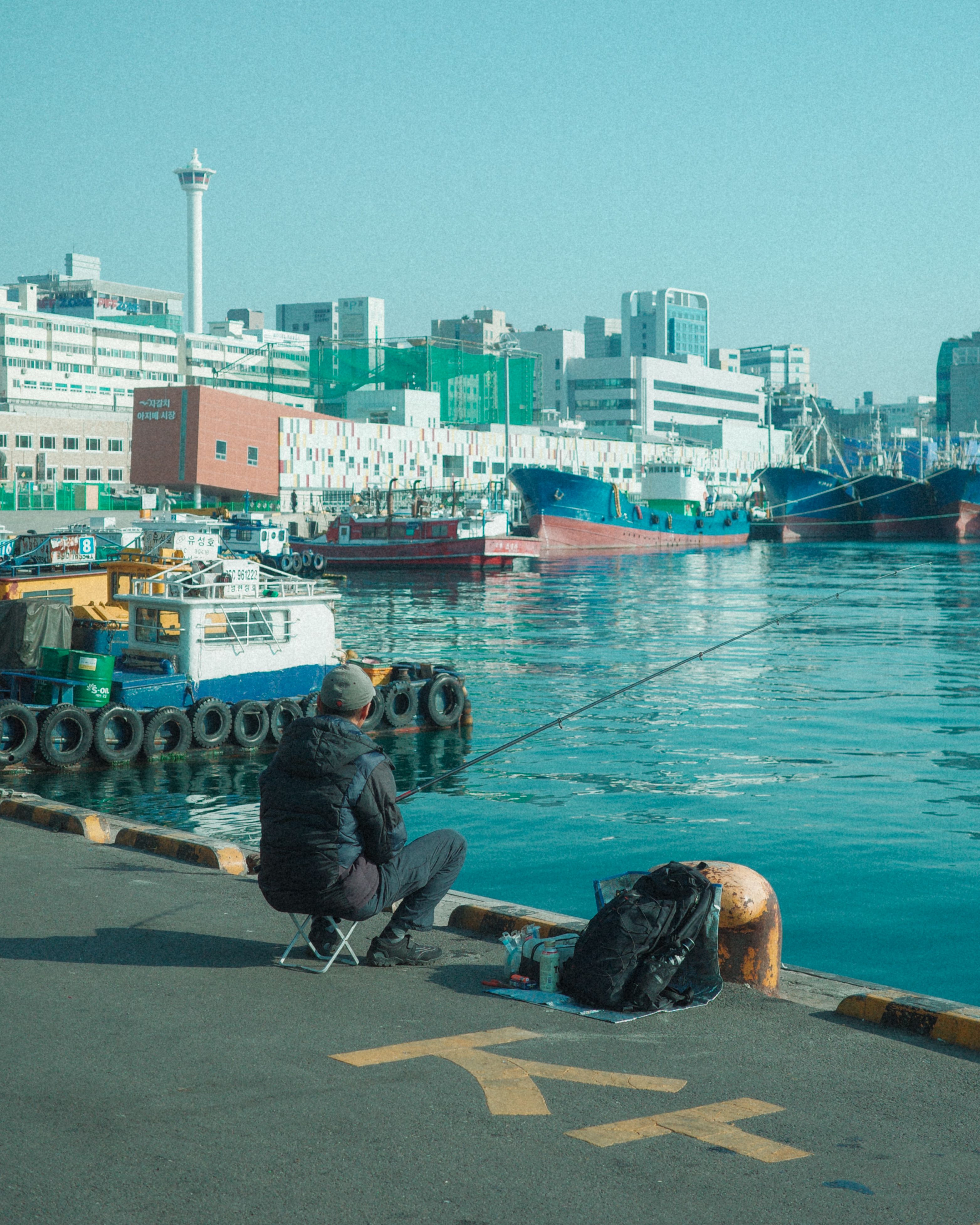 A man fishing at the harbor in Busan, overlooking ships and the city skyline.