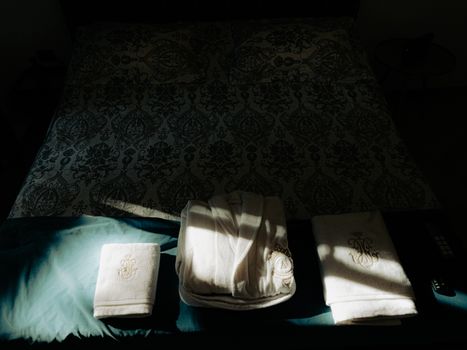 Cozy hotel bedroom scene with patterned bedspread and embroidered towels in soft chiaroscuro lighting.