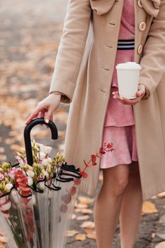 A stylish woman holds a coffee and an umbrella with flowers on a leafy path.