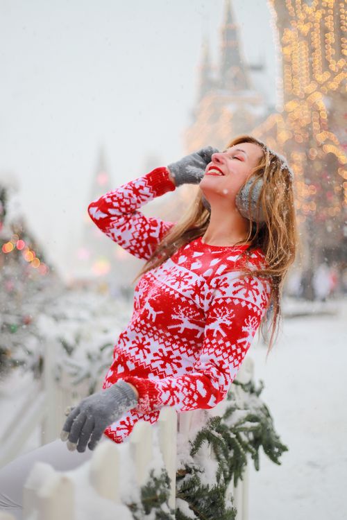 Free Smiling woman wearing a red patterned sweater amidst festive winter snow with holiday lights. Stock Photo