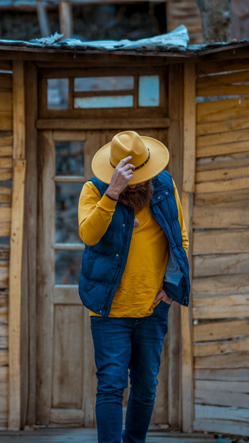 Free Fashionable man in yellow hat and vest walking outdoors by wooden structure during winter. Urban style at its finest. Stock Photo