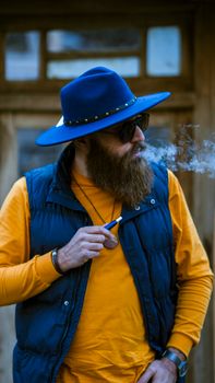 Portrait of a stylish bearded man in sunglasses and blue hat smoking outdoors.