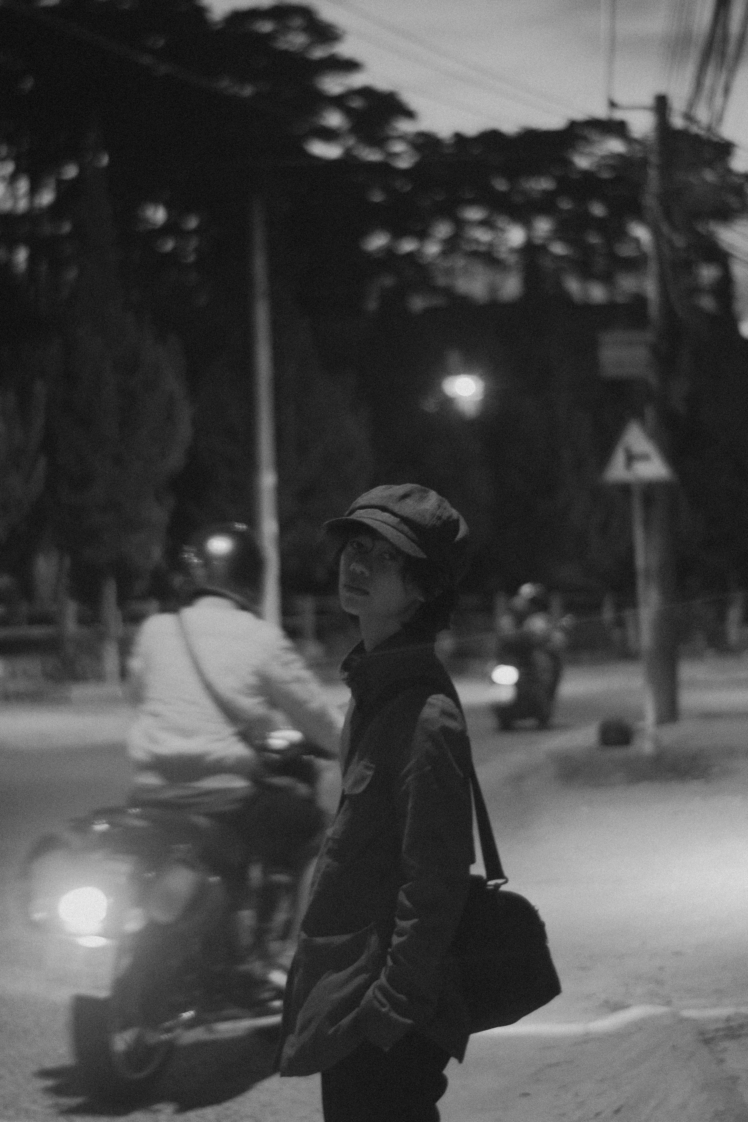 Moody black and white photo of a person standing by a city street at night as motorcycles pass by.