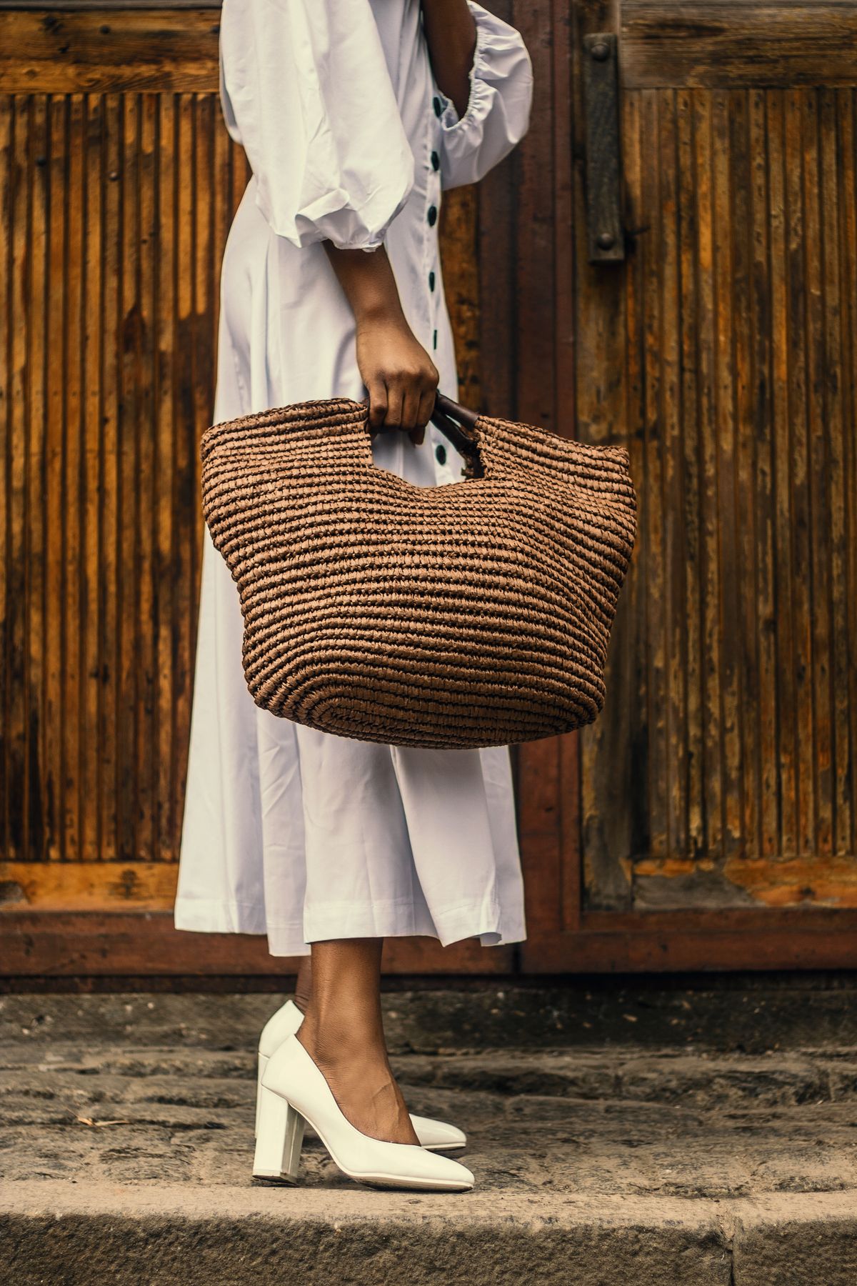 Woman in a tan coat holding a brown raffia bag against a wooden door