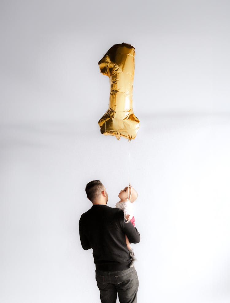 Father Holding His Little Daughter And A Balloon In The Shape Of Number 1 