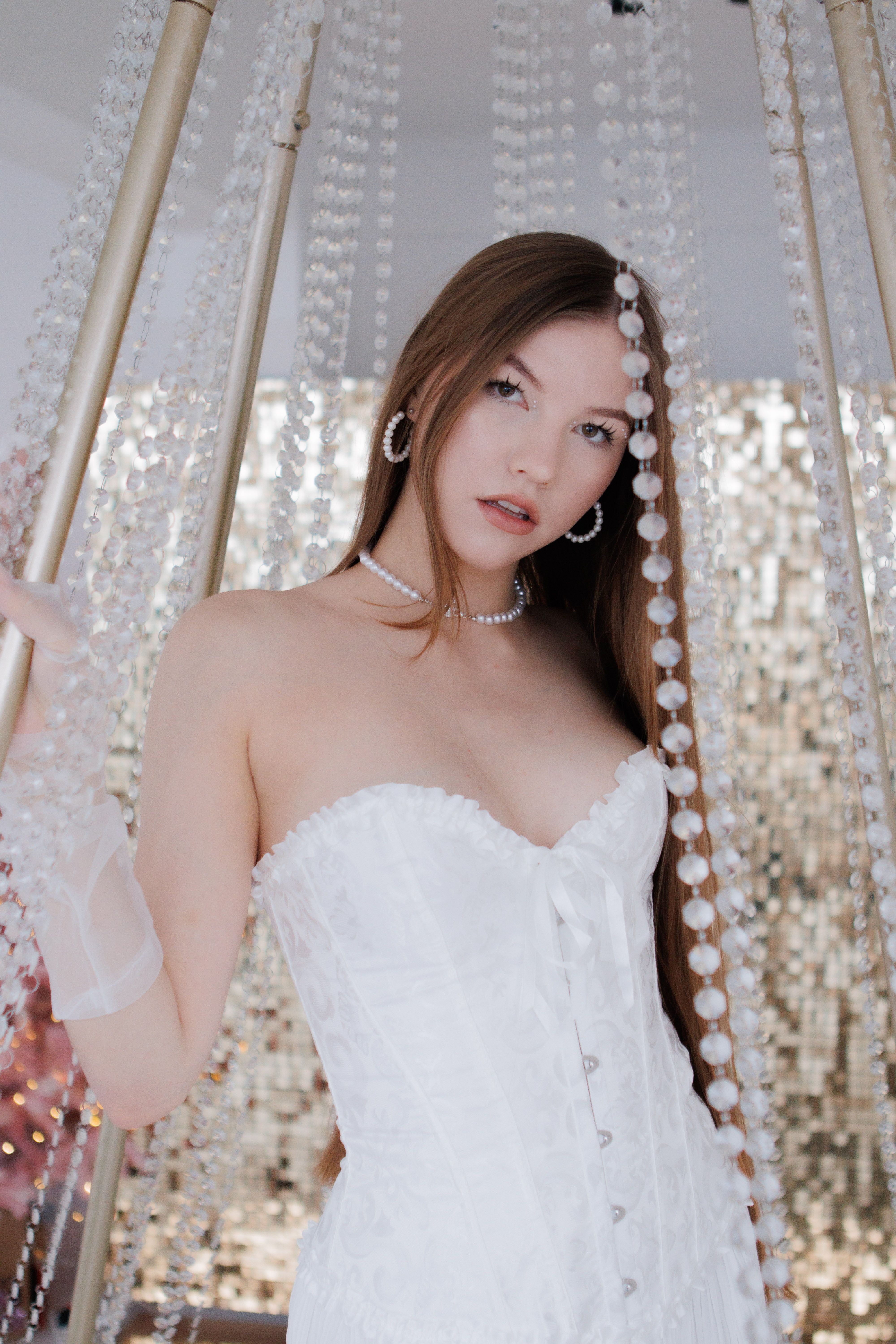 A young woman posing in a white corset amidst hanging bead decorations in a studio setting.