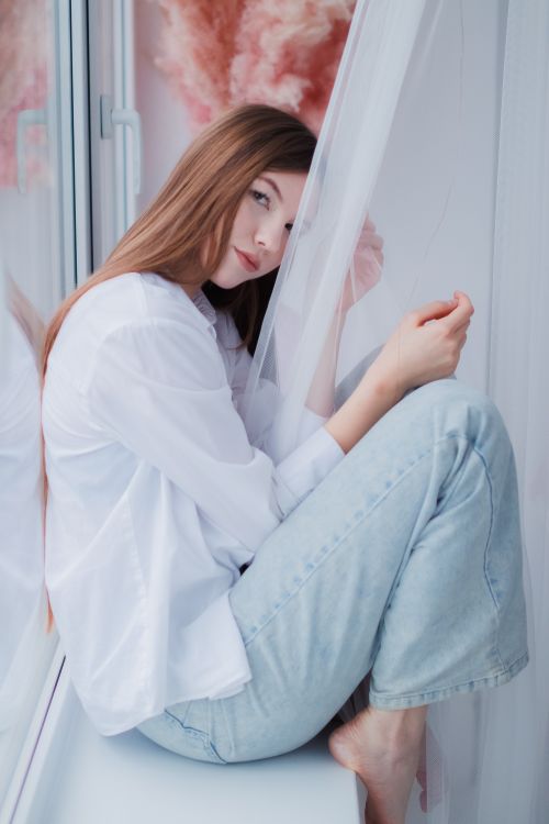 Free Young woman in casual attire sitting on a windowsill, lost in thought, with soft natural lighting. Stock Photo