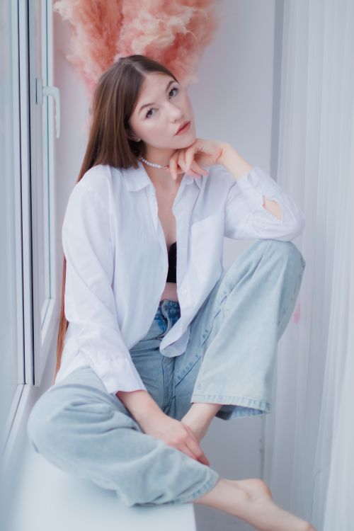 Free A young woman in casual attire sits comfortably on a windowsill in a bright indoor setting. Stock Photo