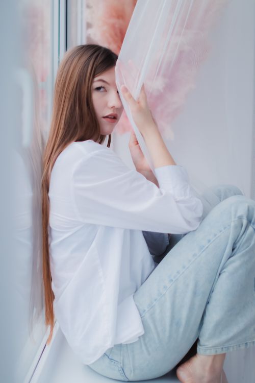 Free Woman with long hair sits on a windowsill, wearing a white shirt and jeans, embracing natural light. Stock Photo