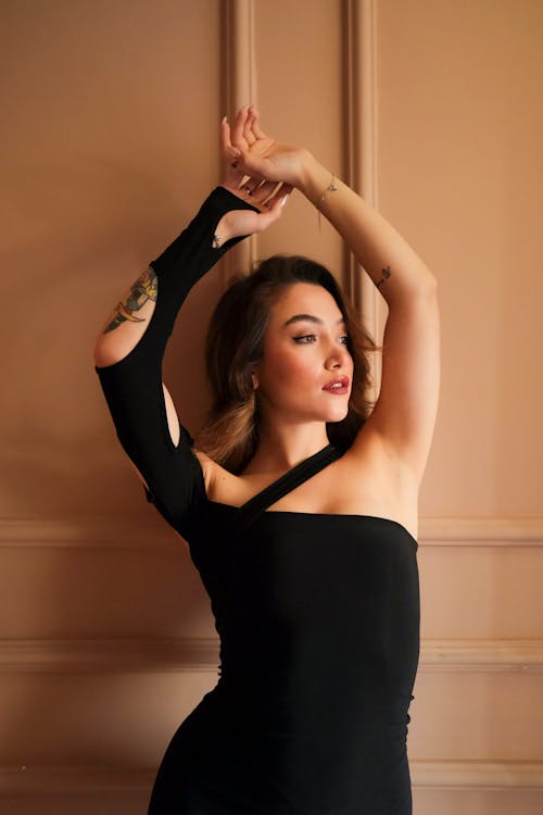 Woman with Tattoos Standing with Arms Raised in Black Dress