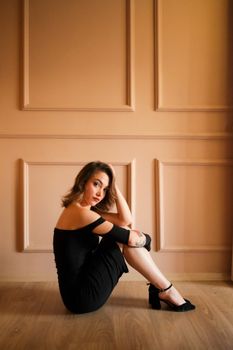 A fashionable woman in a stylish black dress sitting gracefully on the floor indoors.