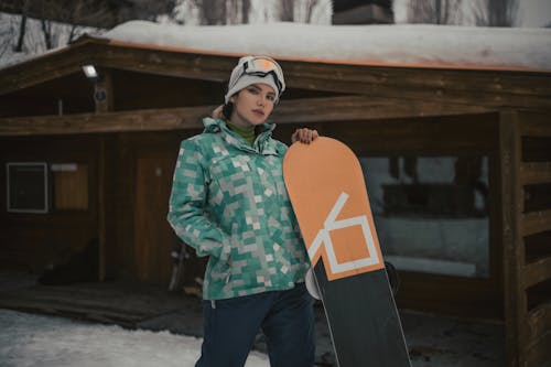 Young Woman Standing with a Snowboard on the Background of a Wooden Hut 